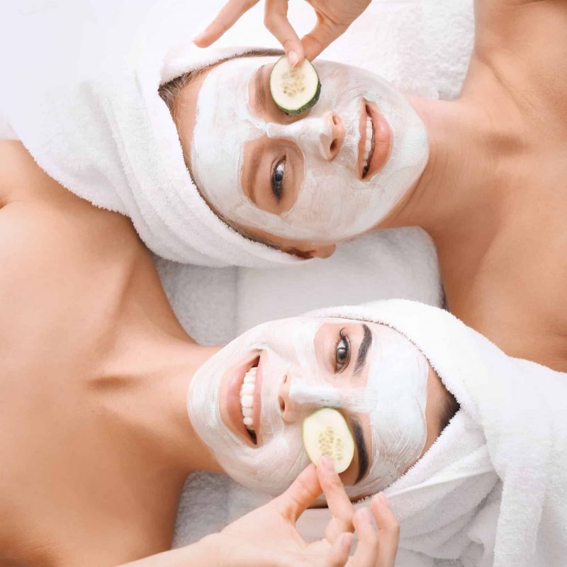 Two women with towels on their heads and beauty product on their faces following an esthetics treatment.
